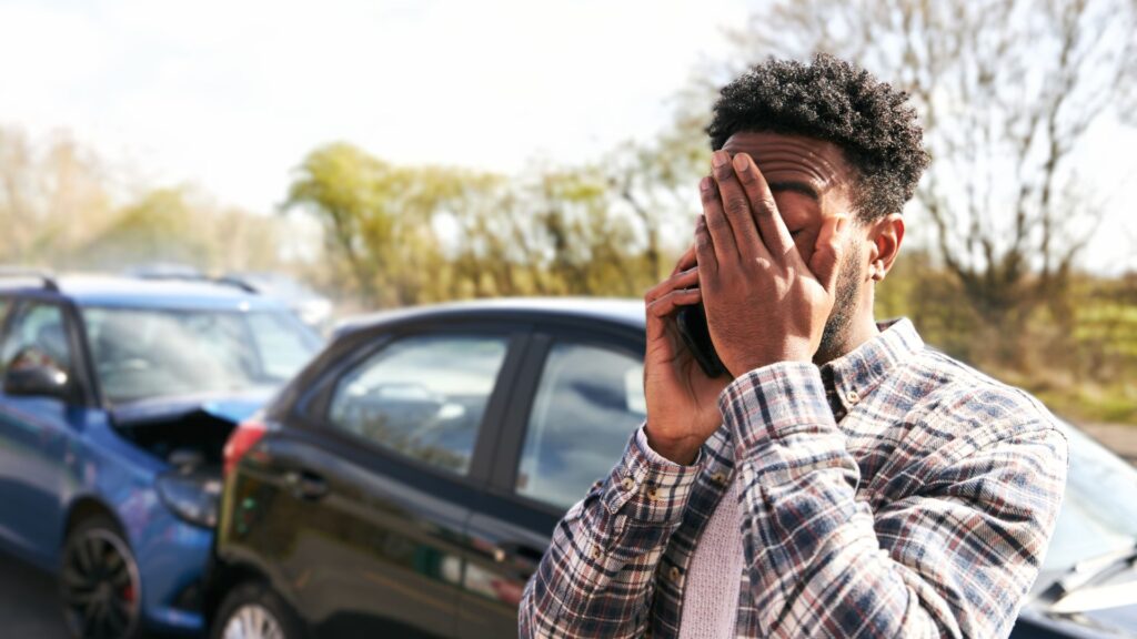Man with hand on his face and cell phone to his ear, standing in front of a car collision.