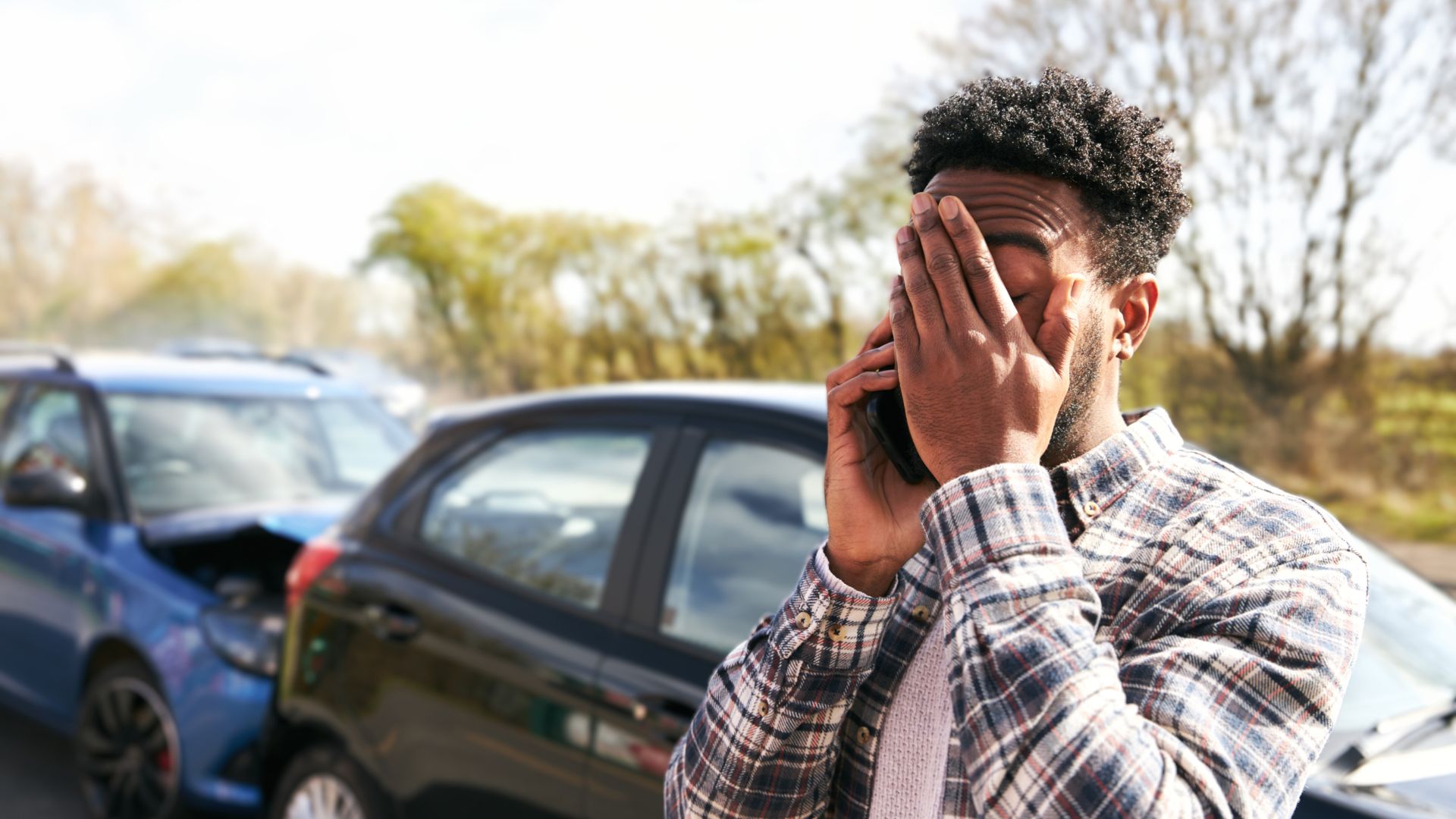 Man with hand on his face and cell phone to his ear, standing in front of a car collision.