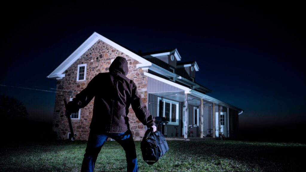 Burglar standing outside a home at night with a duffel bag and crowbar in hand.