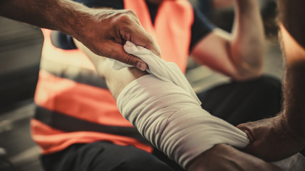 A construction worker getting a burn injury wrapped in gauze by another person.