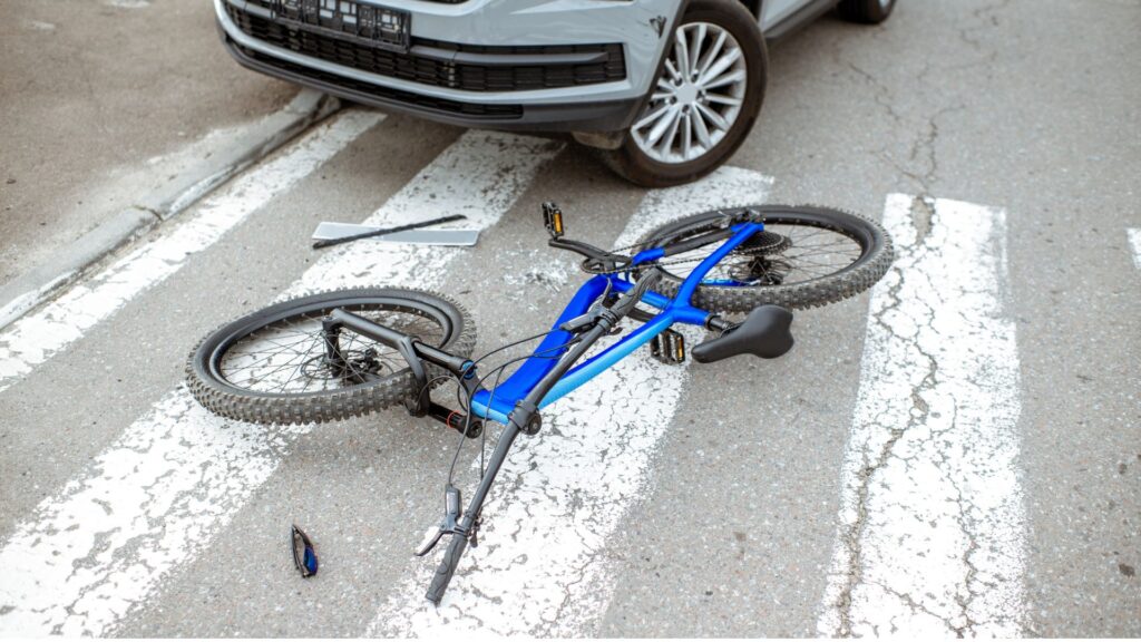 A blue bike fallen on the road with a car behind it.