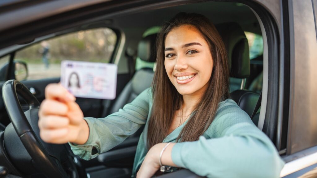 A smiling young woman proudy holding her driver's license in her hand while she's in her car.