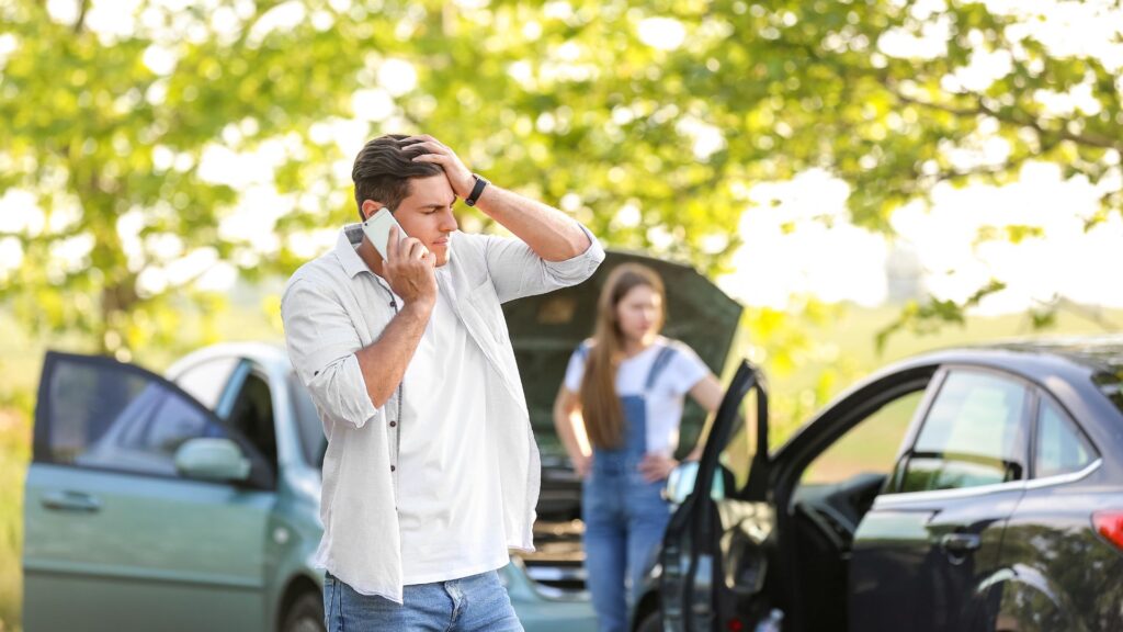 A man on the phone after a car accident and woman looking at the severity of the car crash.