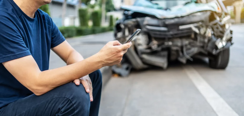 A man sitting on the curb while on his phone after a car accident.