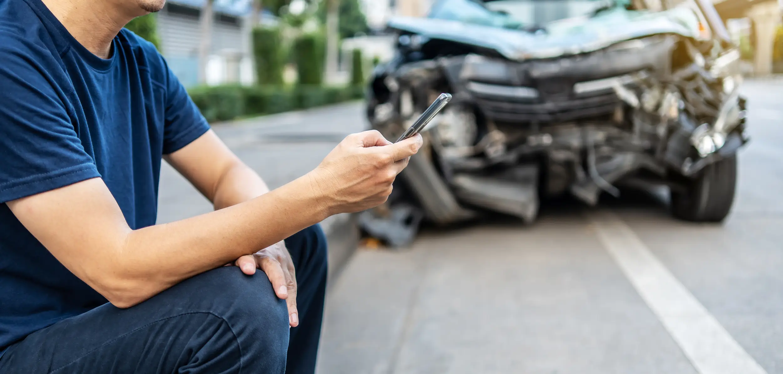 A man sitting on the curb while on his phone after a car accident.