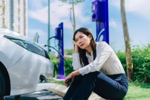 A woman sitting by her electric car while its charging.
