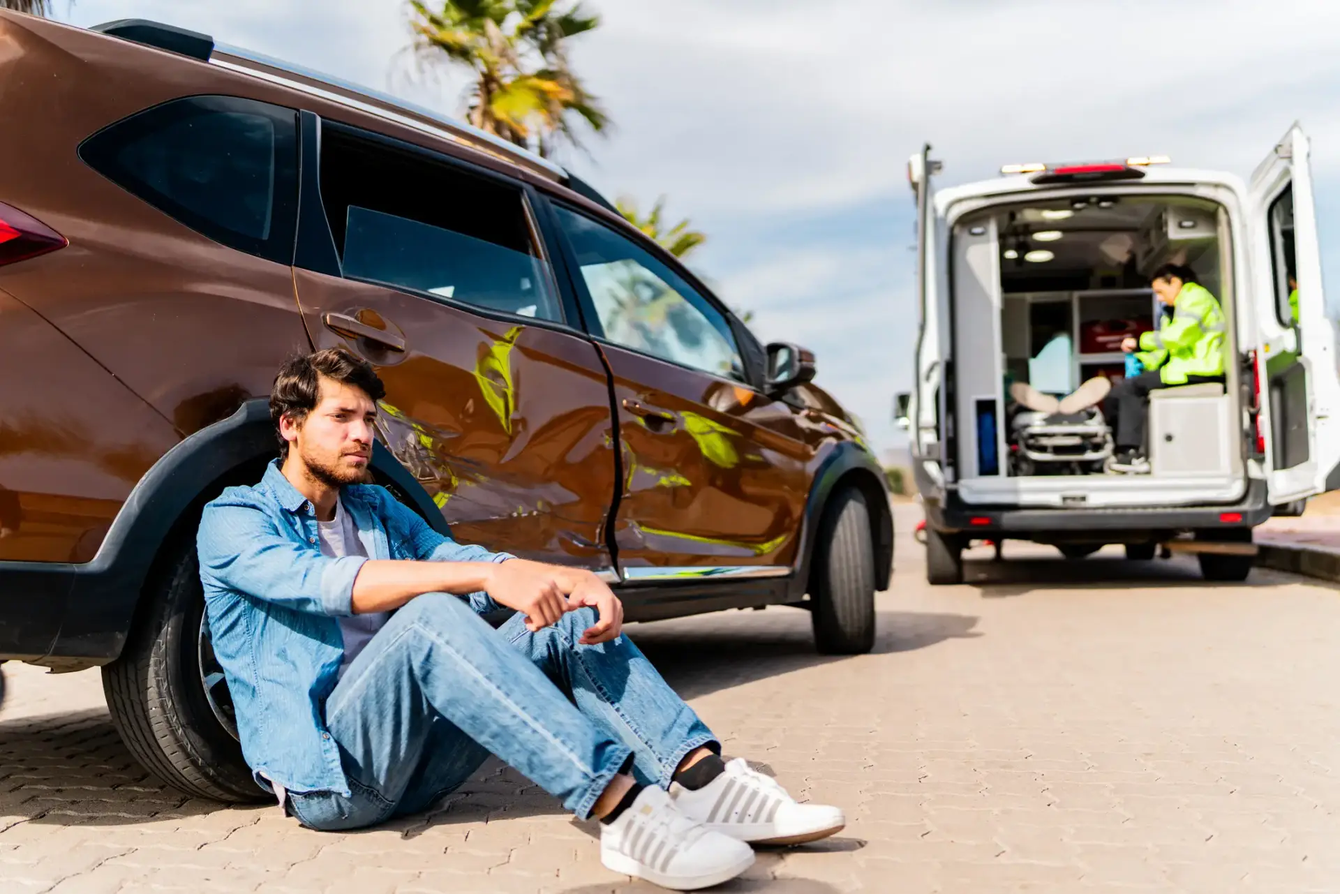 A man sitting by his car after a serious car accident where another person in the ambulance.