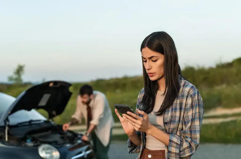 A woman on a phone while a man is trying to fix a broken car on the side of a highway.