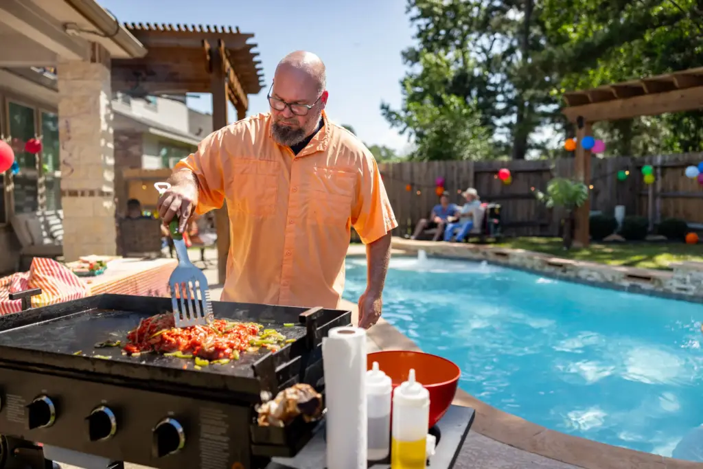 A man cooking on a grill at a family gathering while his family is around the pool.
