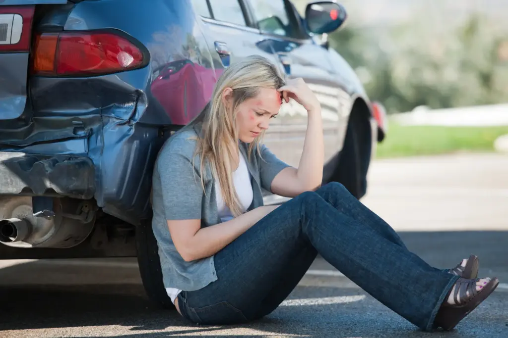 An injured woman sitting and leaning on her car after an accident.
