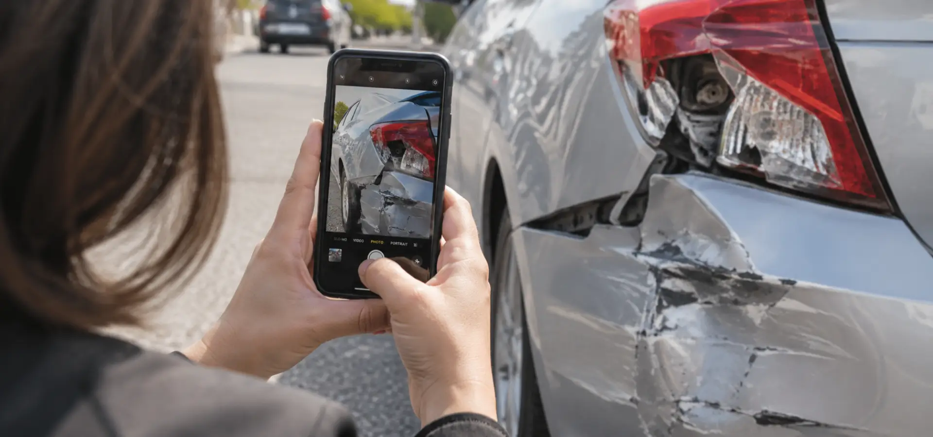 A close-up of a woman using her phone to photograph a dented bumper and broken taillight